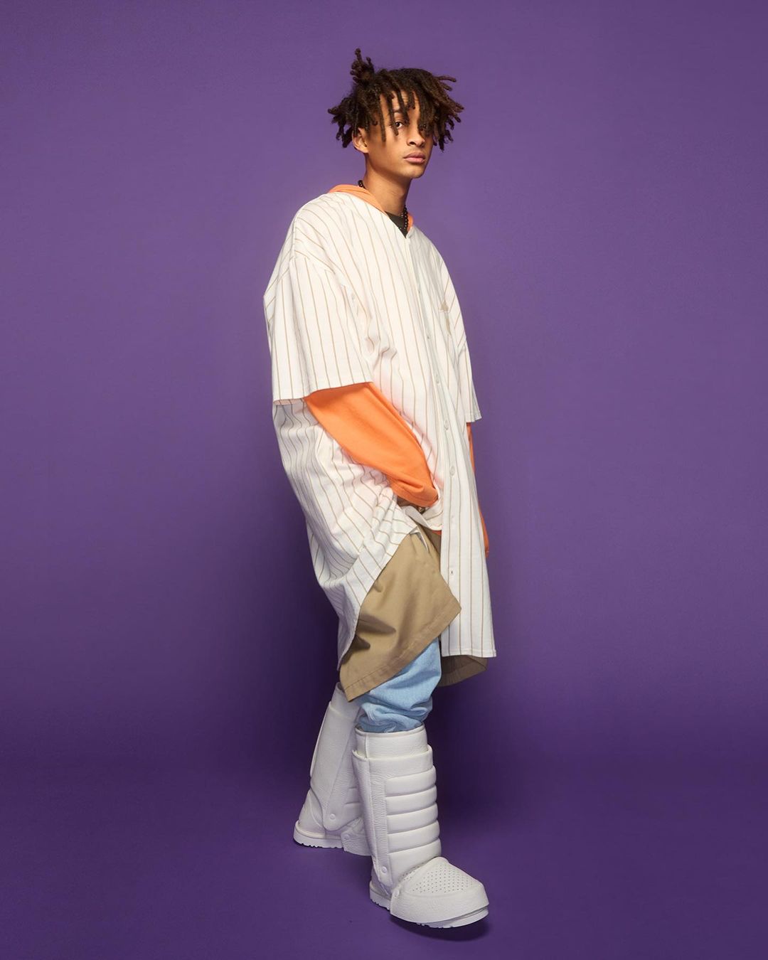 Young adult male in oversized fashion posing against a purple backdrop, wearing a baseball shirt, orange top, khaki pants, and padded boots.