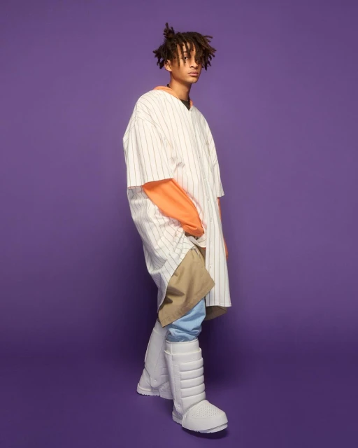 Young adult male in oversized fashion posing against a purple backdrop, wearing a baseball shirt, orange top, khaki pants, and padded boots.