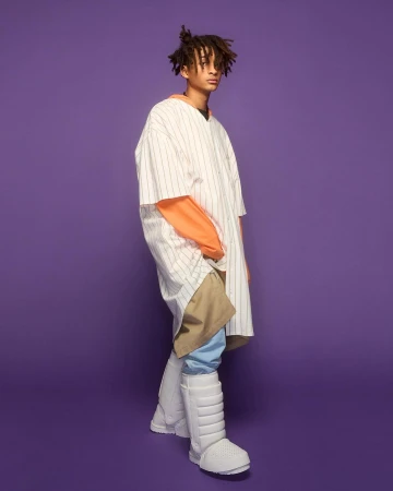 Young adult male in oversized fashion posing against a purple backdrop, wearing a baseball shirt, orange top, khaki pants, and padded boots.
