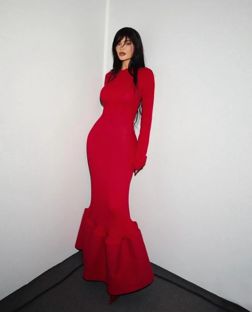 A woman with long dark hair poses in a long-sleeved, floor-length red gown featuring a flared skirt, set against a white wall.