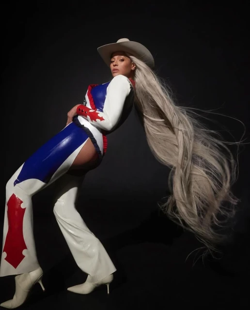 Beyoncé poses in a blue and white outfit with a cowboy hat, and extra-long blonde hair against a dark background in a studio shot.