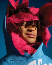Close-up portrait of a man wearing a blue hooded sweatshirt framed by bright pink fabric petals, photographed by Blair Caldwell.