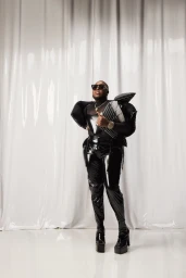 Man wearing dark sunglasses, black structured top, and shiny pants posing in front of a white glossy backdrop.