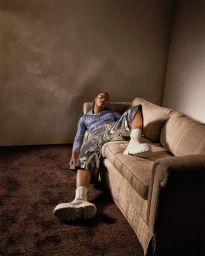 Man reclining on a striped sofa wearing a textured blue shirt, sequined shorts, and chunky white boots.