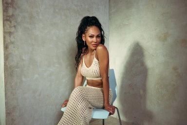 Meagan Good seated on a light blue chair wearing a cream crochet crop top and matching maxi skirt against a textured concrete wall.
