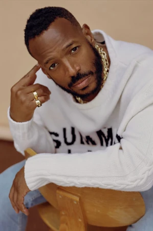 Portrait of Marlon Wayans seated on a wooden chair wearing a white knitted sweater and blue jeans, photographed by Blair Caldwell.