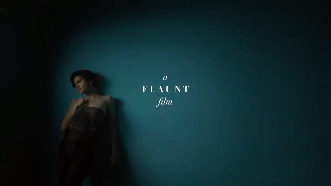 An adult female model with dark hair poses in a brown corset and pants in front of a blue wall with "FLAUNT film" text overlay.