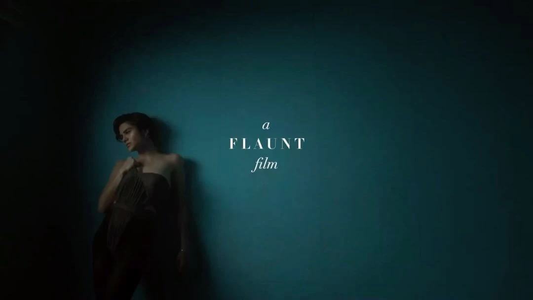 An adult female model with dark hair poses in a brown corset and pants in front of a blue wall with "FLAUNT film" text overlay.