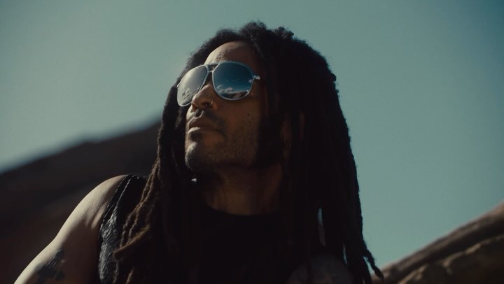 Adult male with dreadlocks and sunglasses looking up toward bright sky on a rooftop. He wears a black tank top and has tattoos.