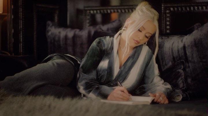 Adult female with blonde hair lying on a couch and writing in a book with a pen in an indoor environment with a dark, moody aesthetic.