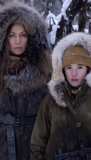 Jennifer Lopez and a child actress in heavy winter clothing and fur-lined hoods, standing in snowy forest.