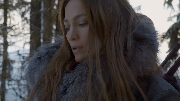 Close-up of adult female with long brown hair wearing a fur-lined hooded coat in a snowy, wooded environment with visible bare trees.