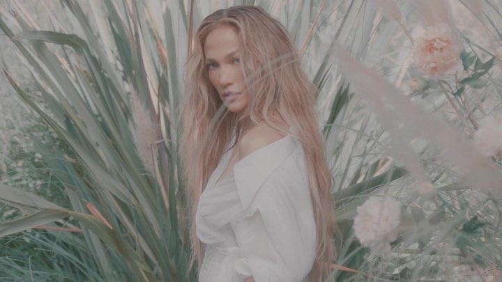 Close-up of adult woman with long blonde hair, wearing a white blouse, posing in front of tall green grasses and pinkish-white flowers.