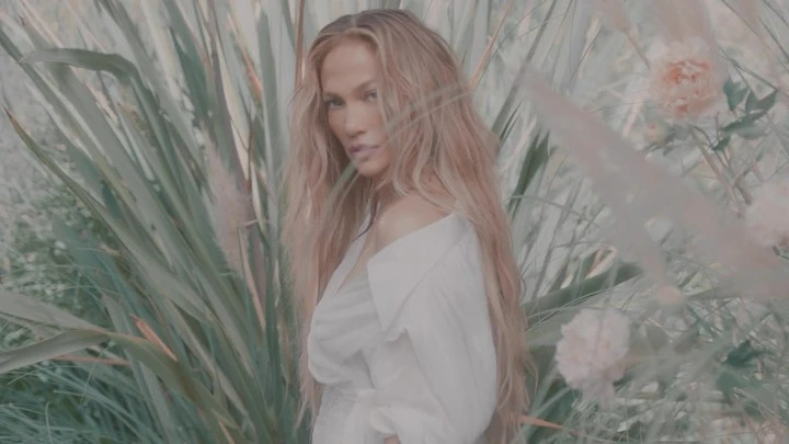 Close-up of adult woman with long blonde hair, wearing a white blouse, posing in front of tall green grasses and pinkish-white flowers.