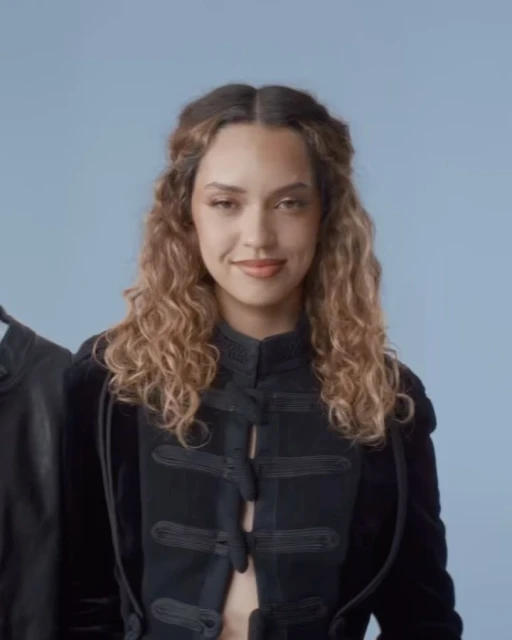 Portrait of a young woman with curly hair wearing a black jacket with embellishments, posing indoors with a light blue backdrop.