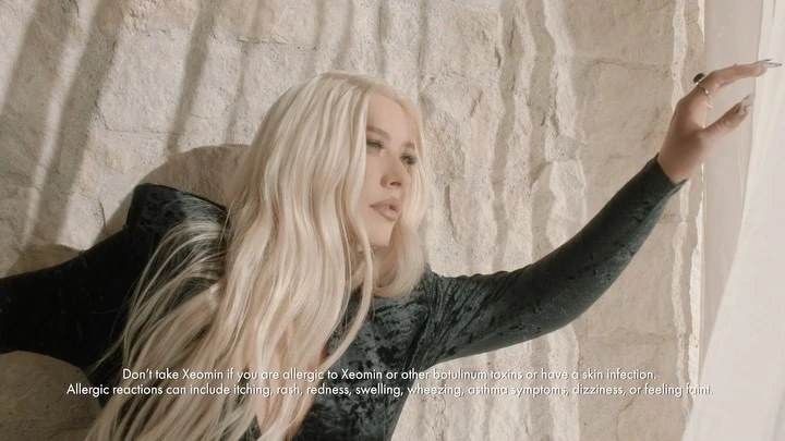 Adult woman with long blonde hair poses in front of a textured stone wall, wearing a dark velvet top, with an extended hand.