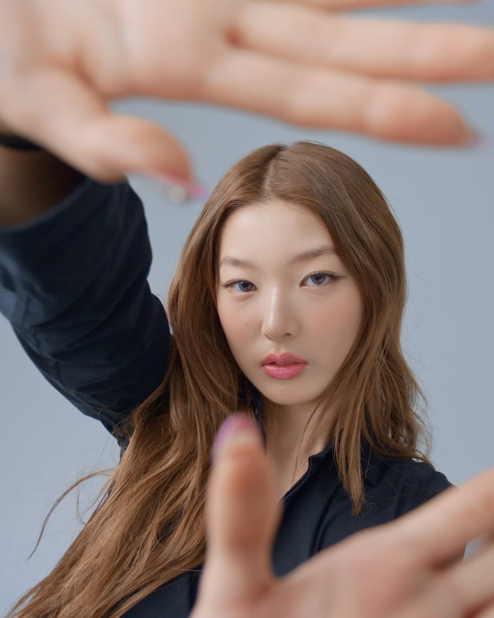 Close-up shot of an adult woman with light brown hair and fair skin, framed by human hands on a neutral grey background in a studio.