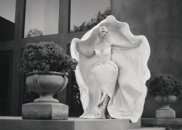 Anya Taylor-Joy wearing a sculptural white gown standing on a stone terrace between two large planters.