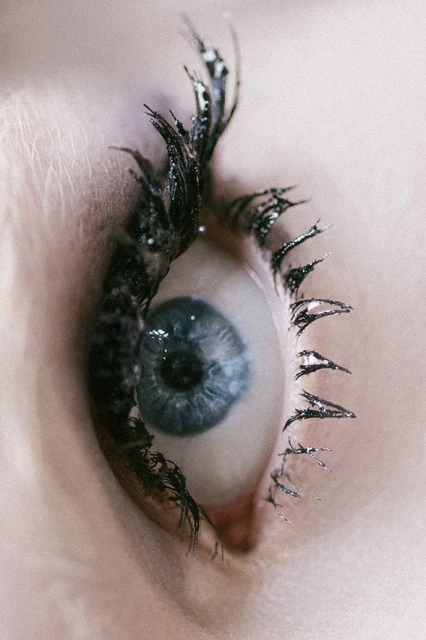 Close-up of a blue eye with heavy black mascara on the eyelashes. The skin around the eye is pale, and the focus is on the eye itself.