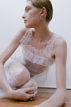 A woman in a white sheer ruffled dress seated on a wooden floor against a plain wall photographed by Erika Astrid