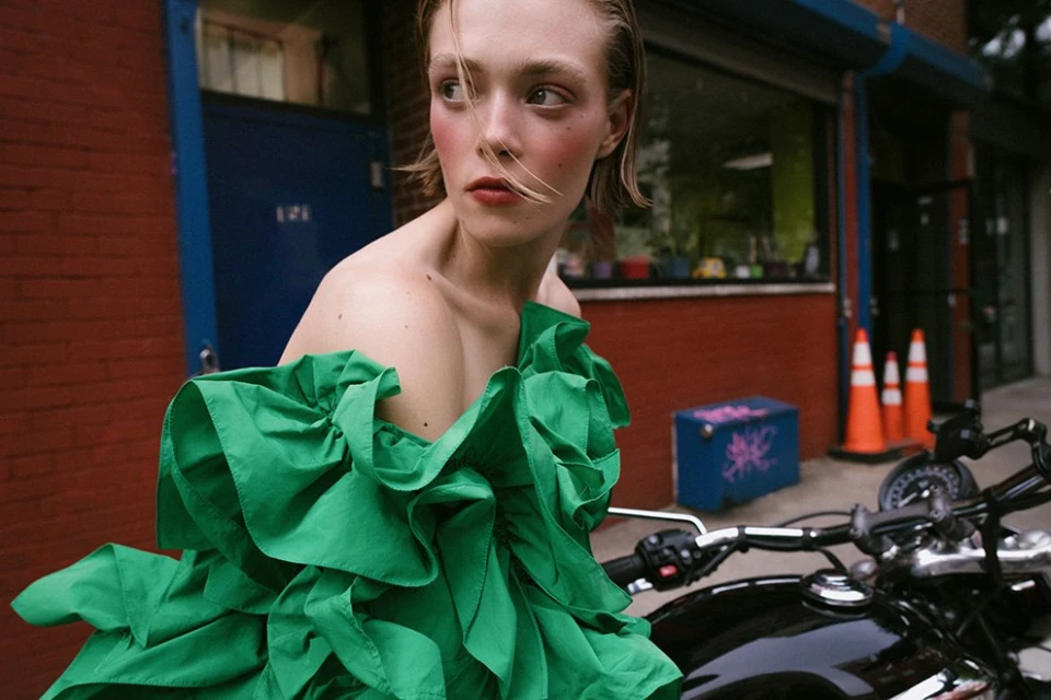 Woman in green dress poses on a motorcycle in an urban setting with brick buildings and greenery, featuring a stylish fashion portrait.