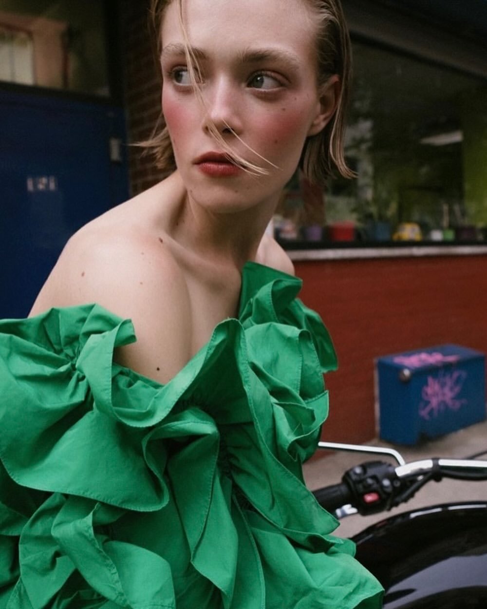Blonde model in green dress poses on a black motorcycle in an urban setting with an expression looking to the side in portrait.  