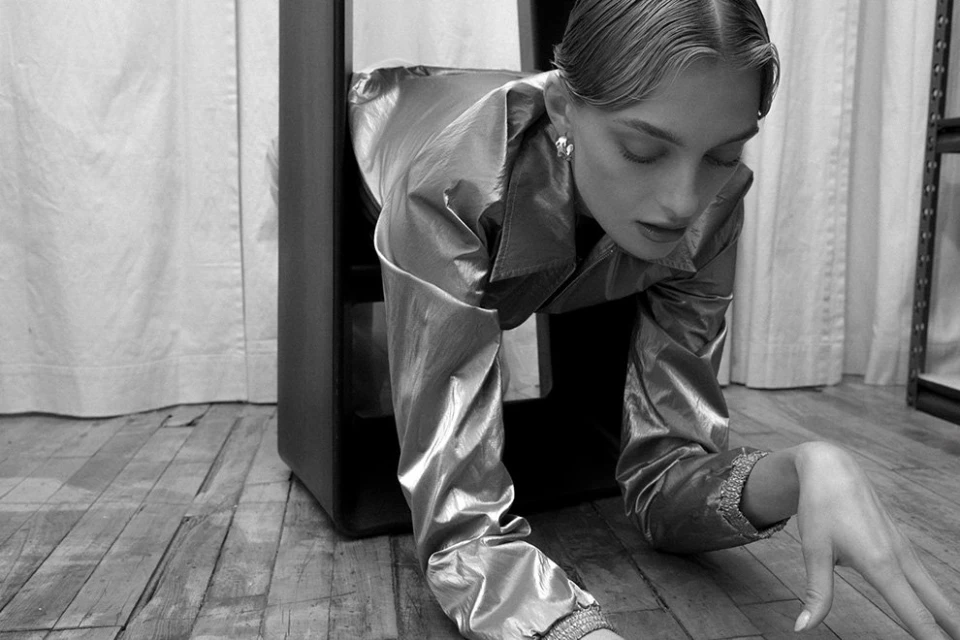 Monochrome shot of a woman in a metallic jacket crawling through a dark, rectangular structure indoors on a wooden floor.