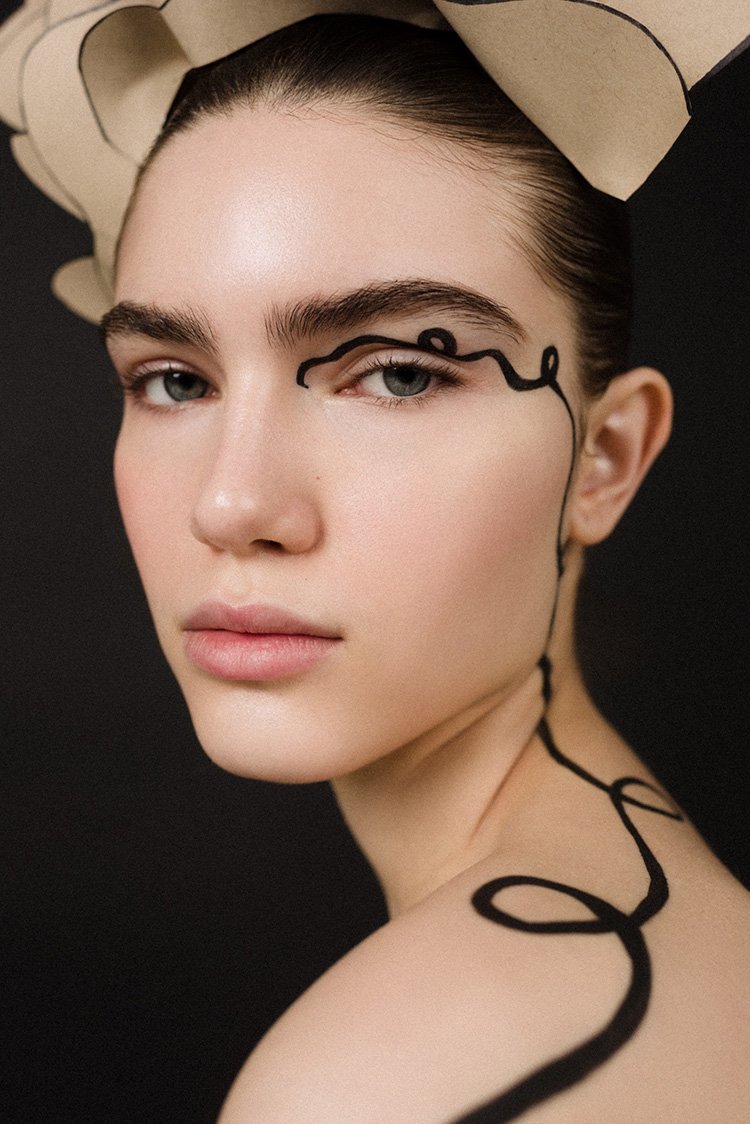Close-up studio shot of a young adult woman with creative black eyeliner and a beige headpiece posing against a dark background.
