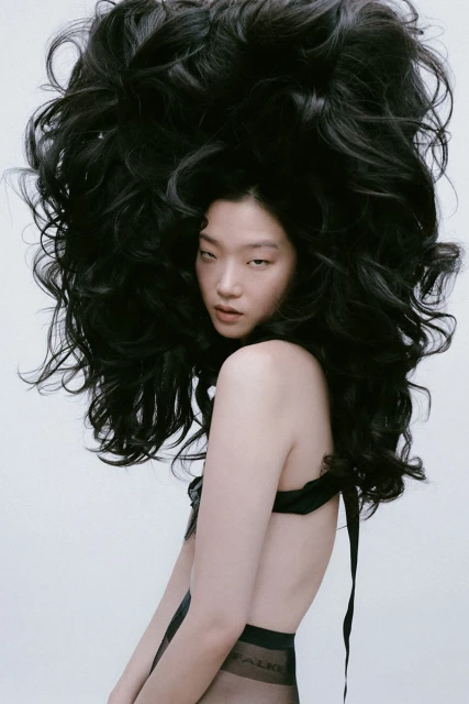 Woman with voluminous, wavy black hair poses in a studio with light skin, sheer hosiery, and a black top with ribbon straps.