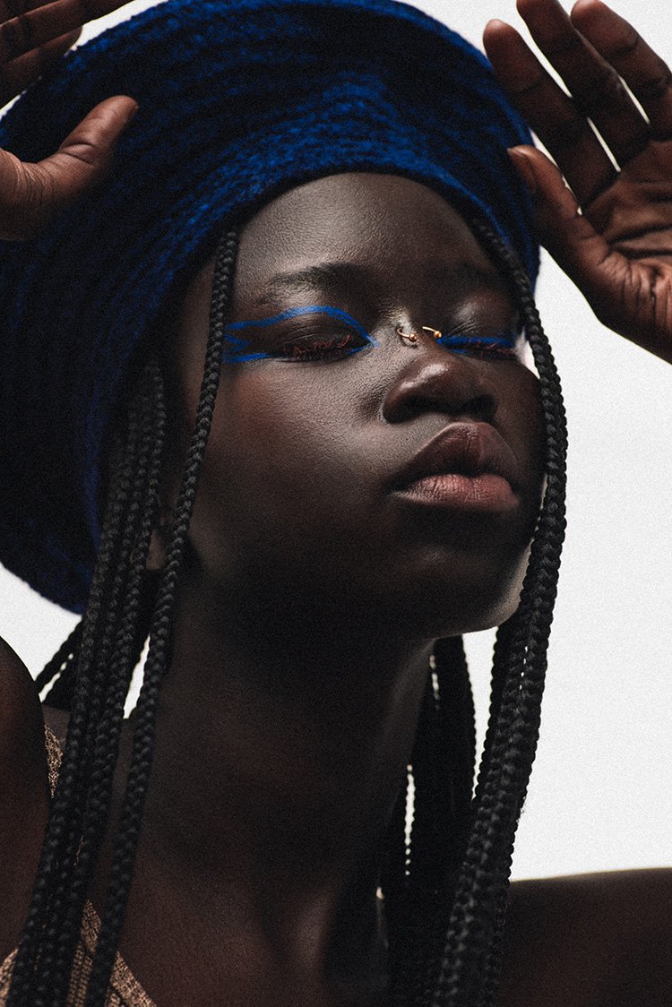Close-up portrait of an African woman with blue eye makeup and a blue textured hat, gazing downward in a brightly lit, simple setting.