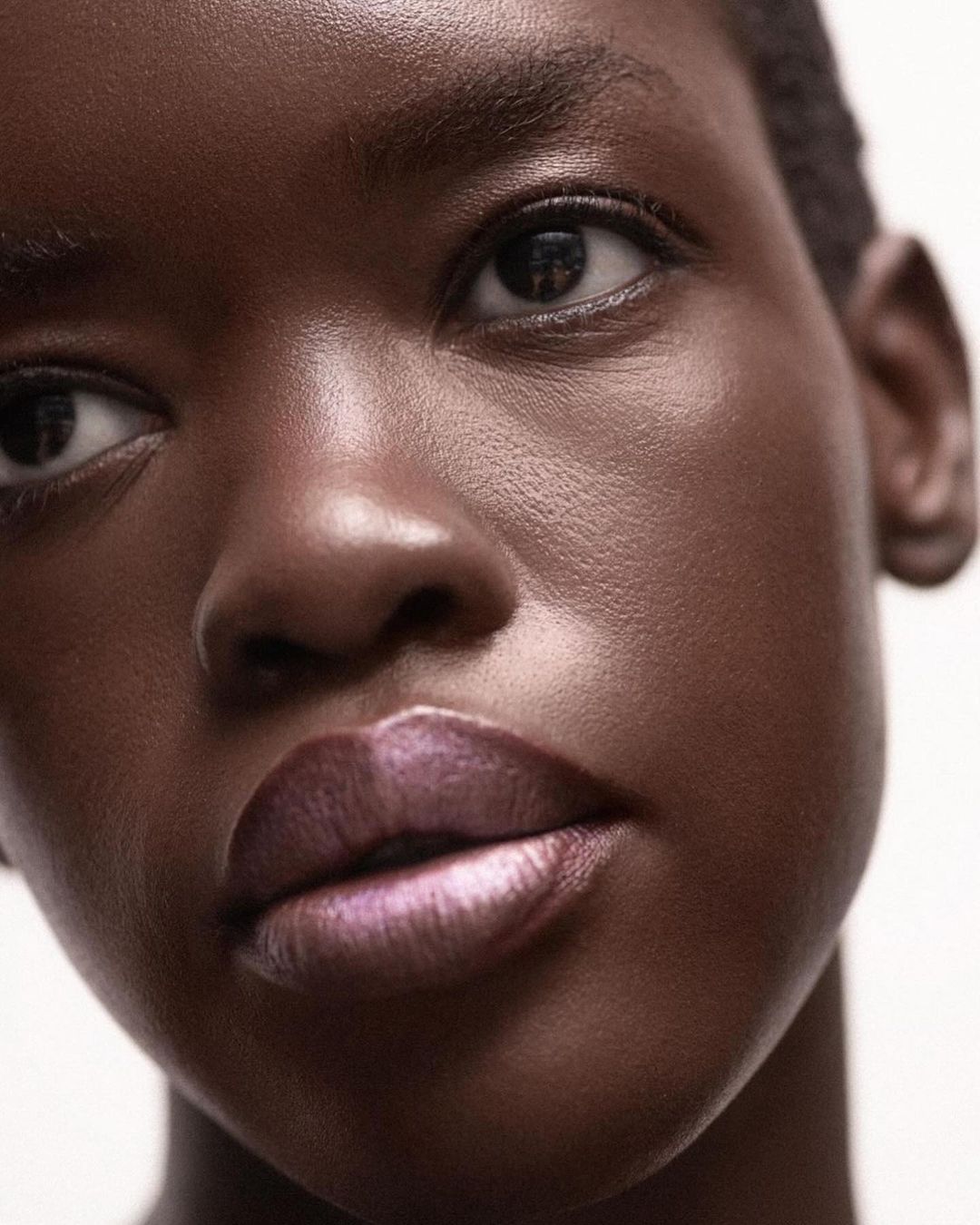 Close-up portrait of a dark-skinned woman with short hair and glossy lips, illuminated by soft, diffused lighting against a neutral backdrop.