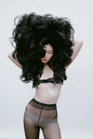 A woman with voluminous long black hair framing her face and body wearing a dark star shaped top and sheer tights against a plain background