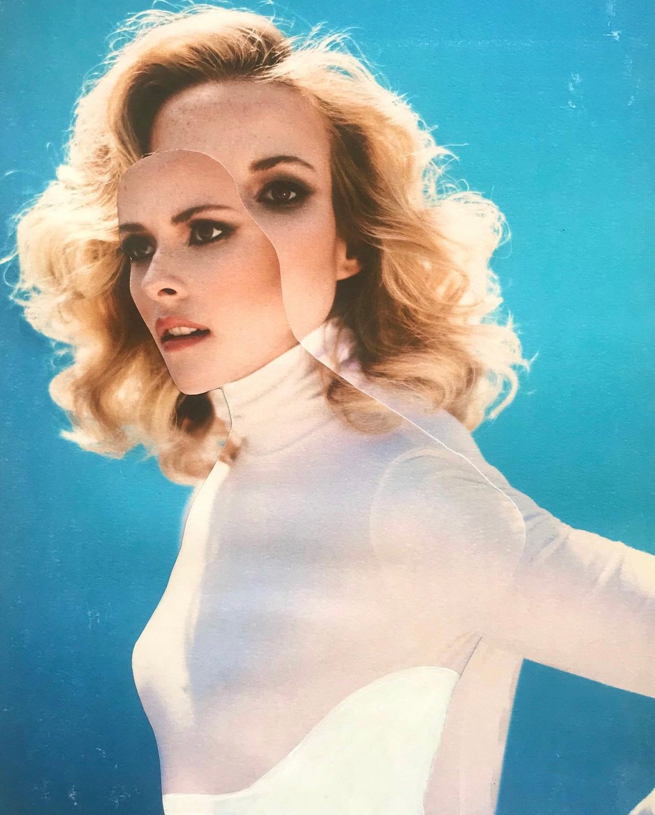 Woman with blonde wavy hair and smoky eye makeup wearing a white turtleneck, posing against a gradient blue background in a cutout style.