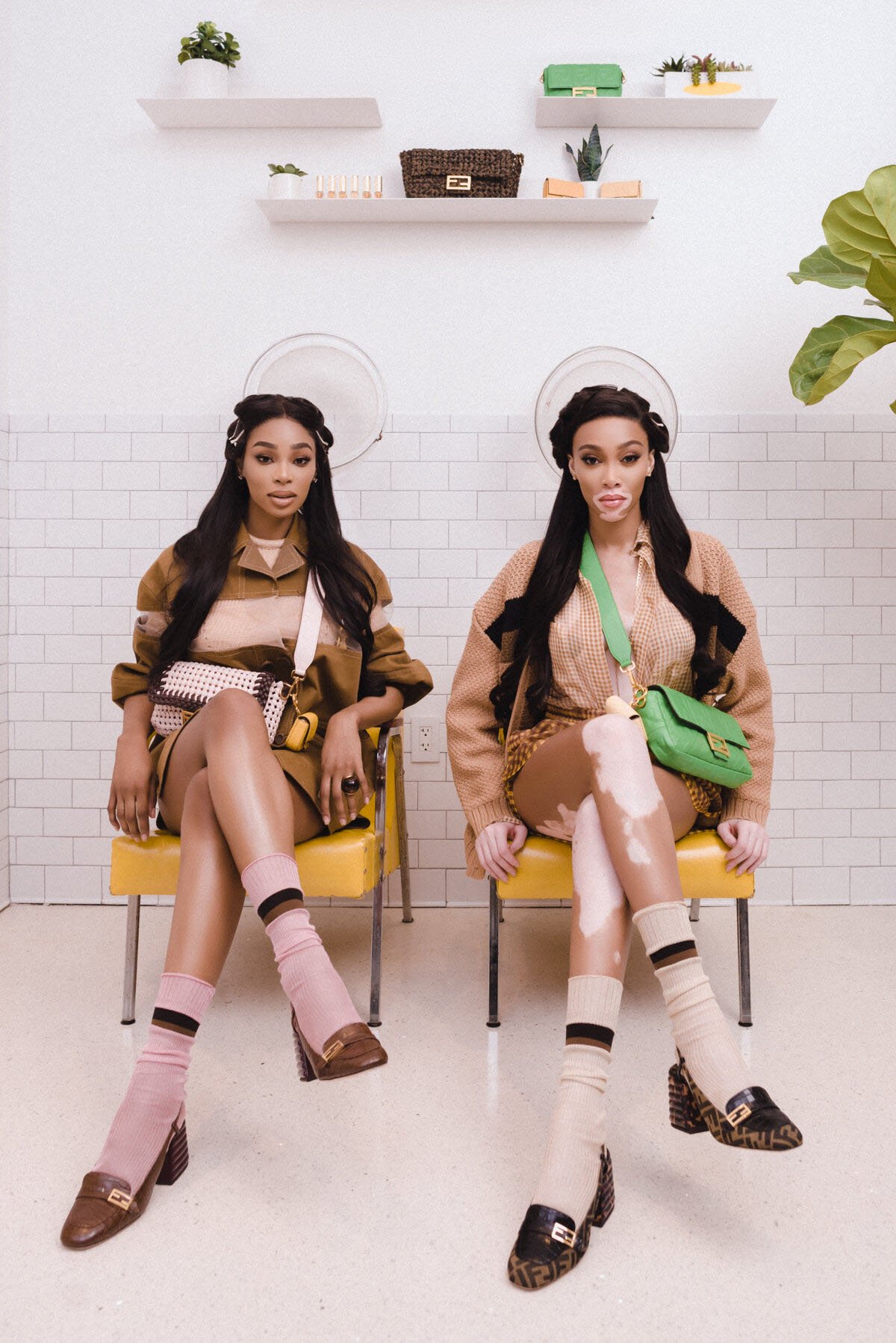 Two women sit in yellow chairs, showcasing Fendi loafers, bags, and outfits in a light-filled salon with white brick walls and shelves.