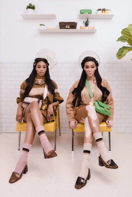 Two women sit in yellow chairs, showcasing Fendi loafers, bags, and outfits in a light-filled salon with white brick walls and shelves.