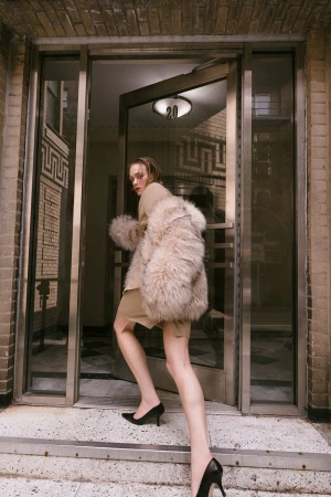 Stylish woman in fur coat enters a building, wearing black high heels on steps, captured in an urban setting with natural lighting.