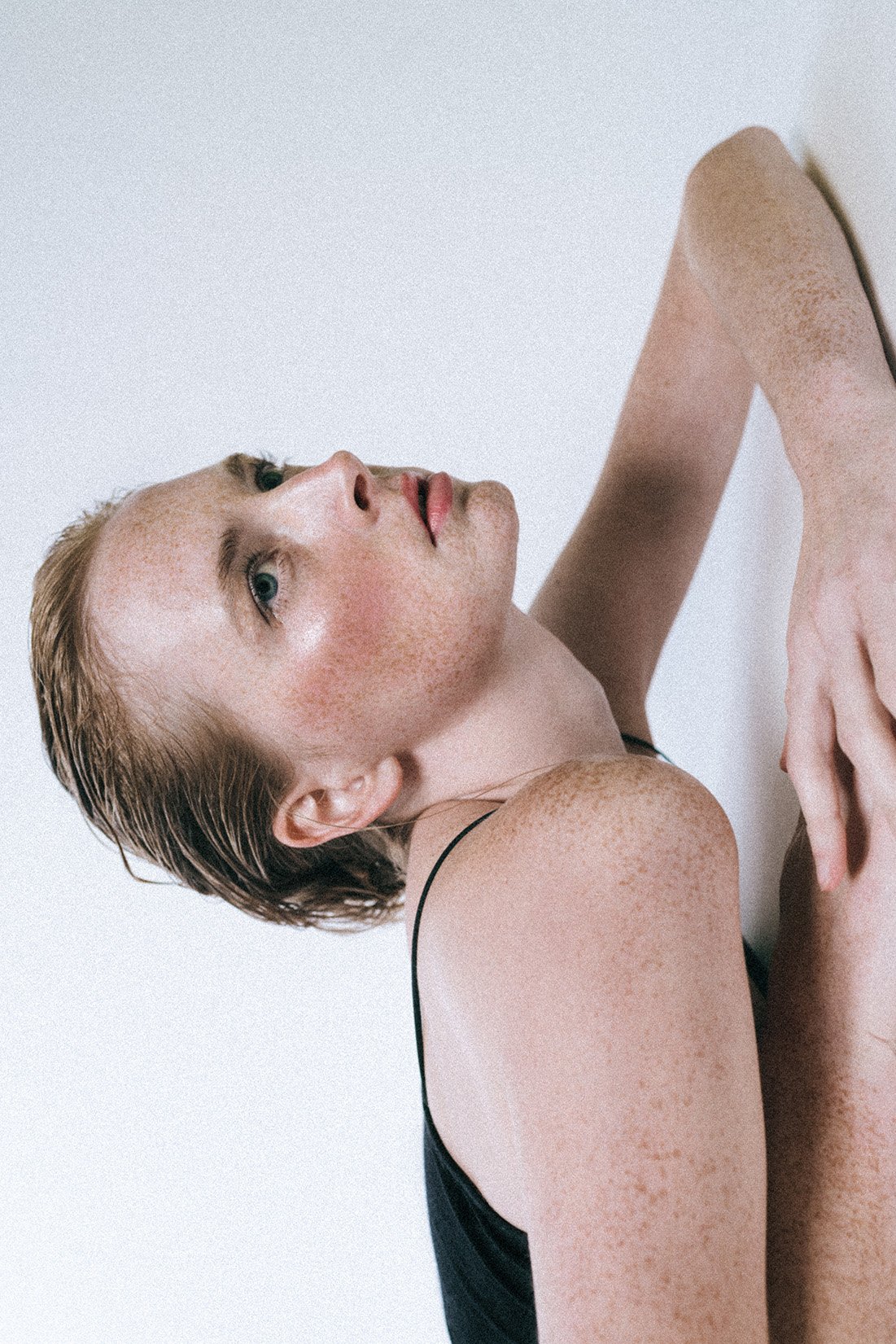 Portrait of a freckled woman with wet hair wearing a black spaghetti strap top against a white wall by Erika Astrid
