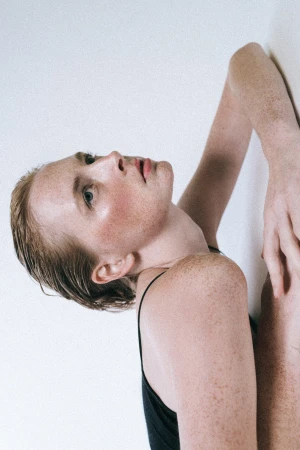 Portrait of a freckled woman with wet hair wearing a black spaghetti strap top against a white wall by Erika Astrid