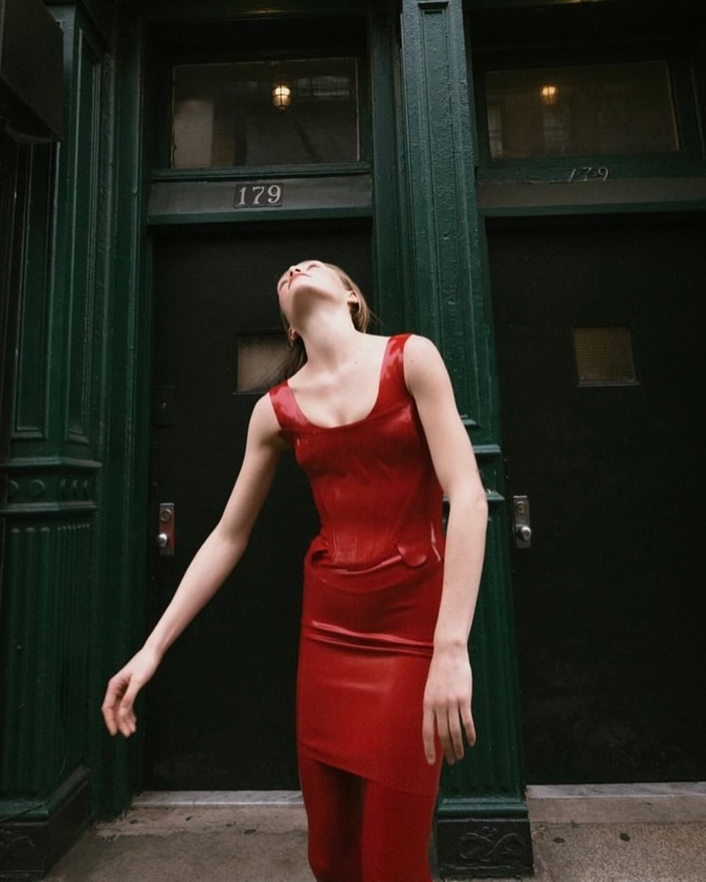 Model wearing a glossy red dress with head tilted back standing in front of dark green double doors.