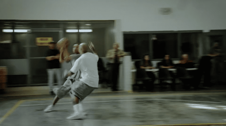 Two adult men playing basketball indoors with onlookers in the background, inside of a basketball court.