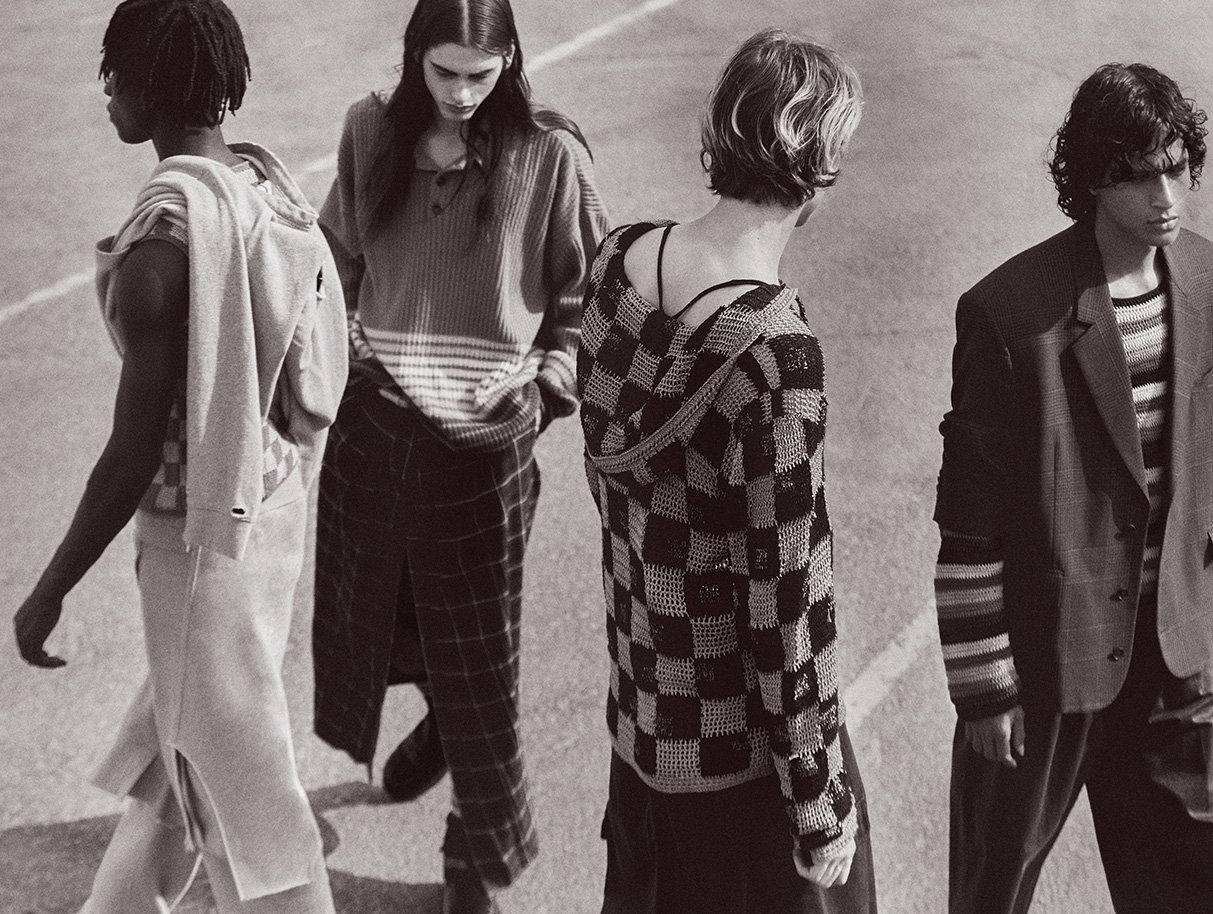 Four fashion models stand shoulder to shoulder on concrete in muted tones. The models wear knitwear and tailored pieces in a muted color palette.