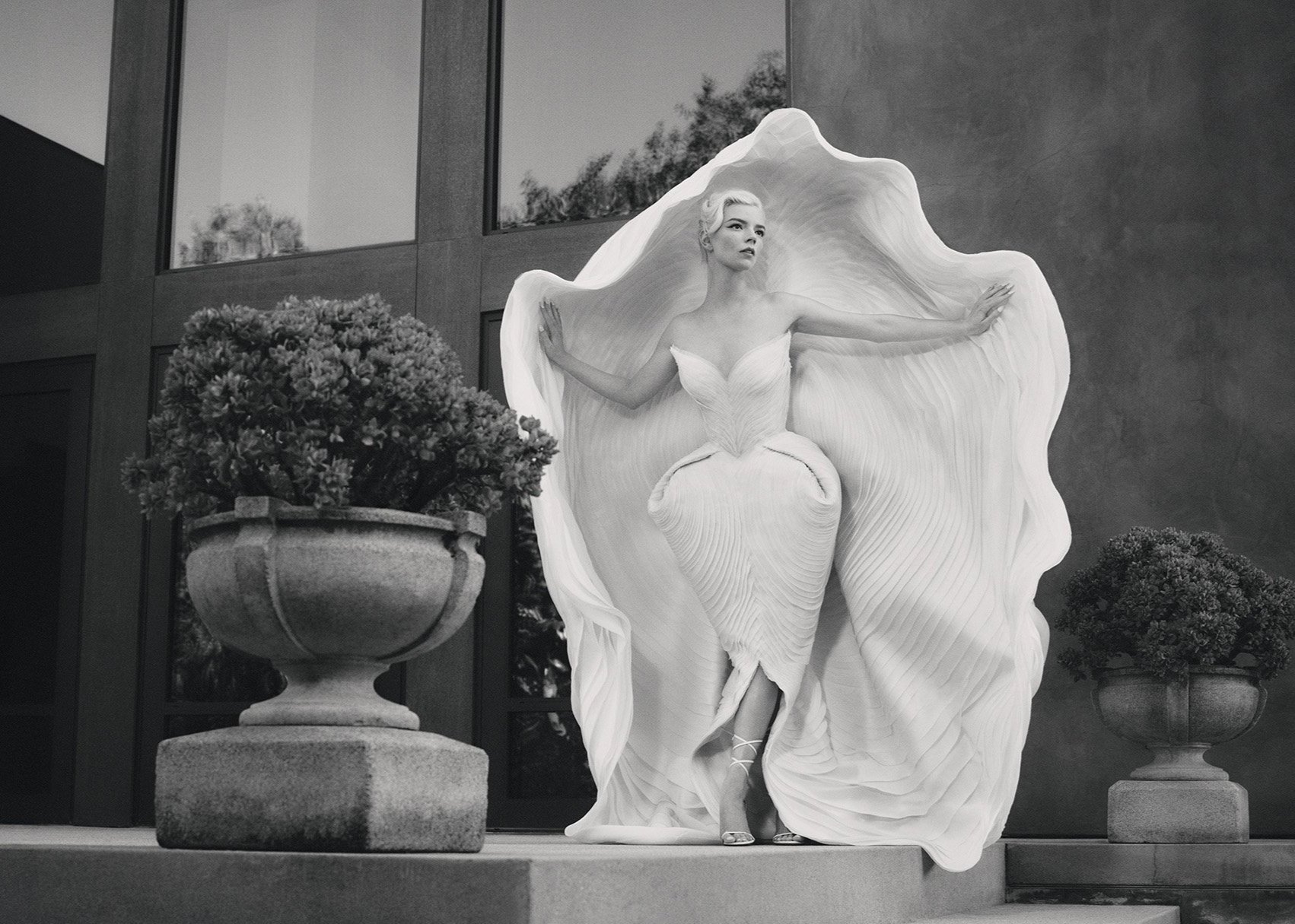 Anya Taylor-Joy poses in a white dress and cape outside a building with large windows and potted plants in a monochrome fashion shot.