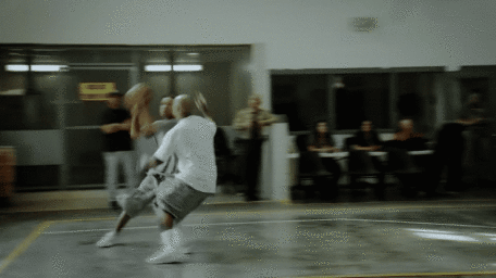 Two men playing basketball inside an indoor gym with spectators watching from the sidelines.