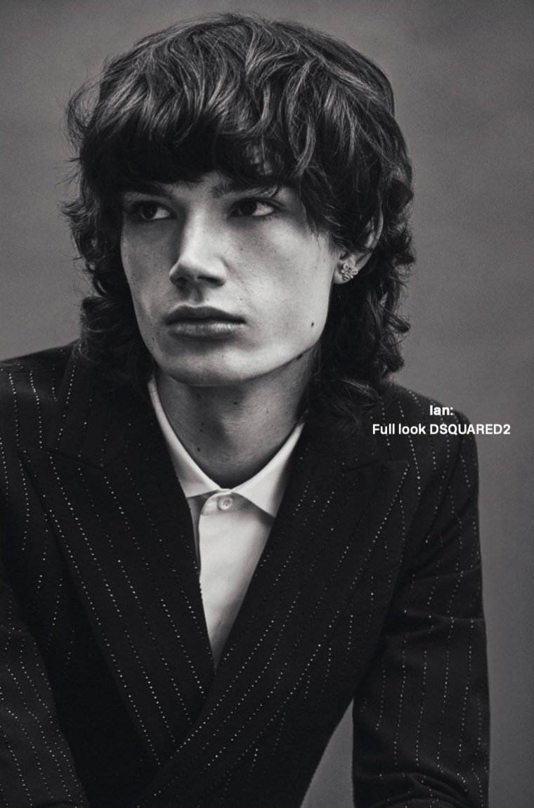 Close-up portrait of a young adult man with wavy dark hair, wearing a pinstripe blazer and white collared shirt, looking off camera.