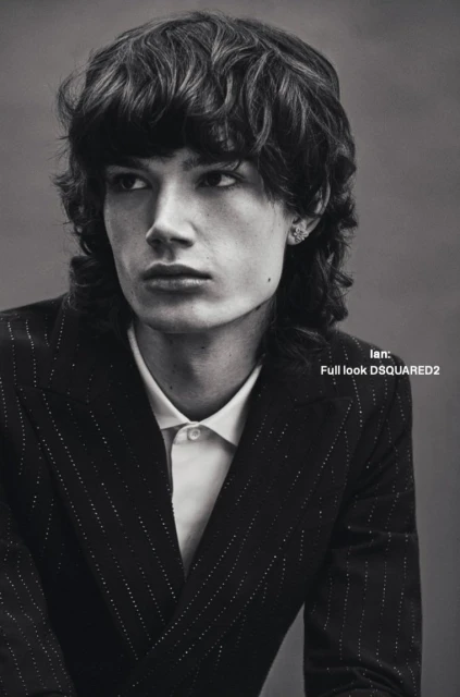 Close-up portrait of a young adult man with wavy dark hair, wearing a pinstripe blazer and white collared shirt, looking off camera.