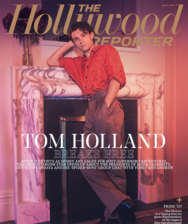 Magazine cover featuring Tom Holland leaning against a marble fireplace, wearing a red shirt and brown pants, indoor with flowers.