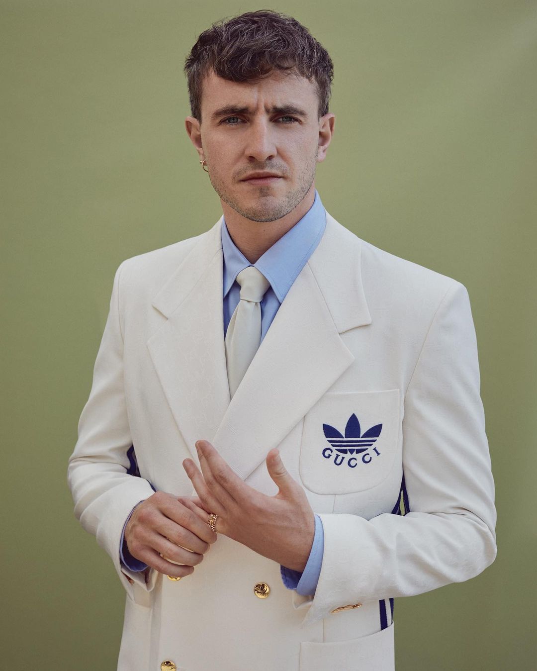 Portrait of adult male in white suit with Gucci logo looking at the camera in a photo studio with a light green backdrop.