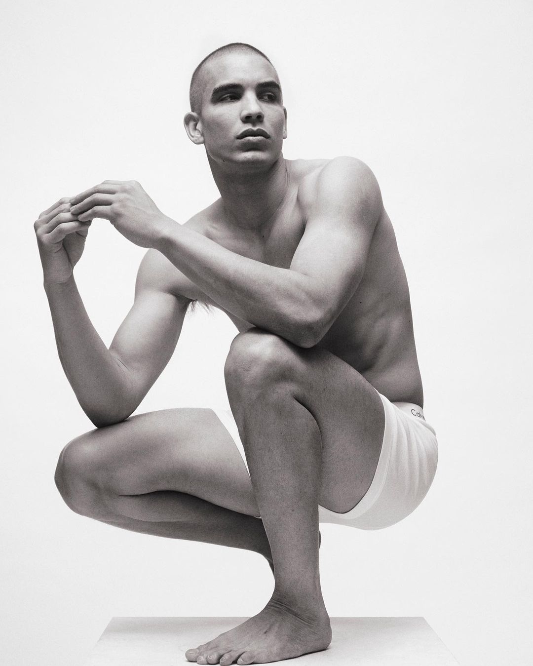 Black and white portrait of a muscular, bald adult male in a crouching pose, wearing white underwear, against a plain white backdrop.