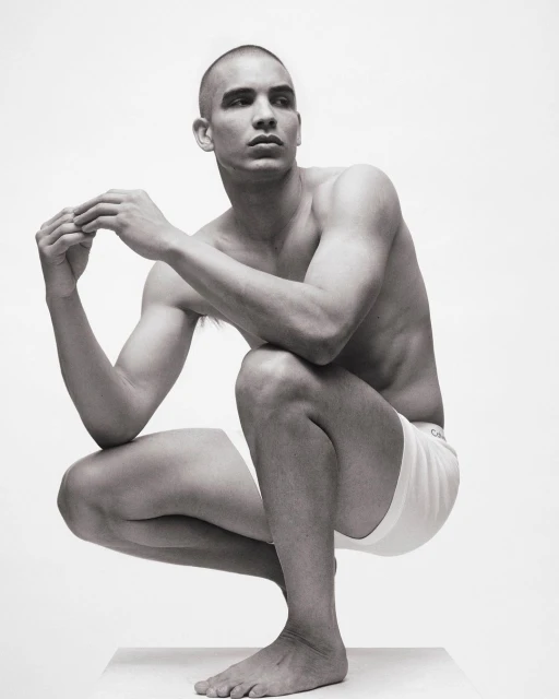 Black and white portrait of a muscular, bald adult male in a crouching pose, wearing white underwear, against a plain white backdrop.