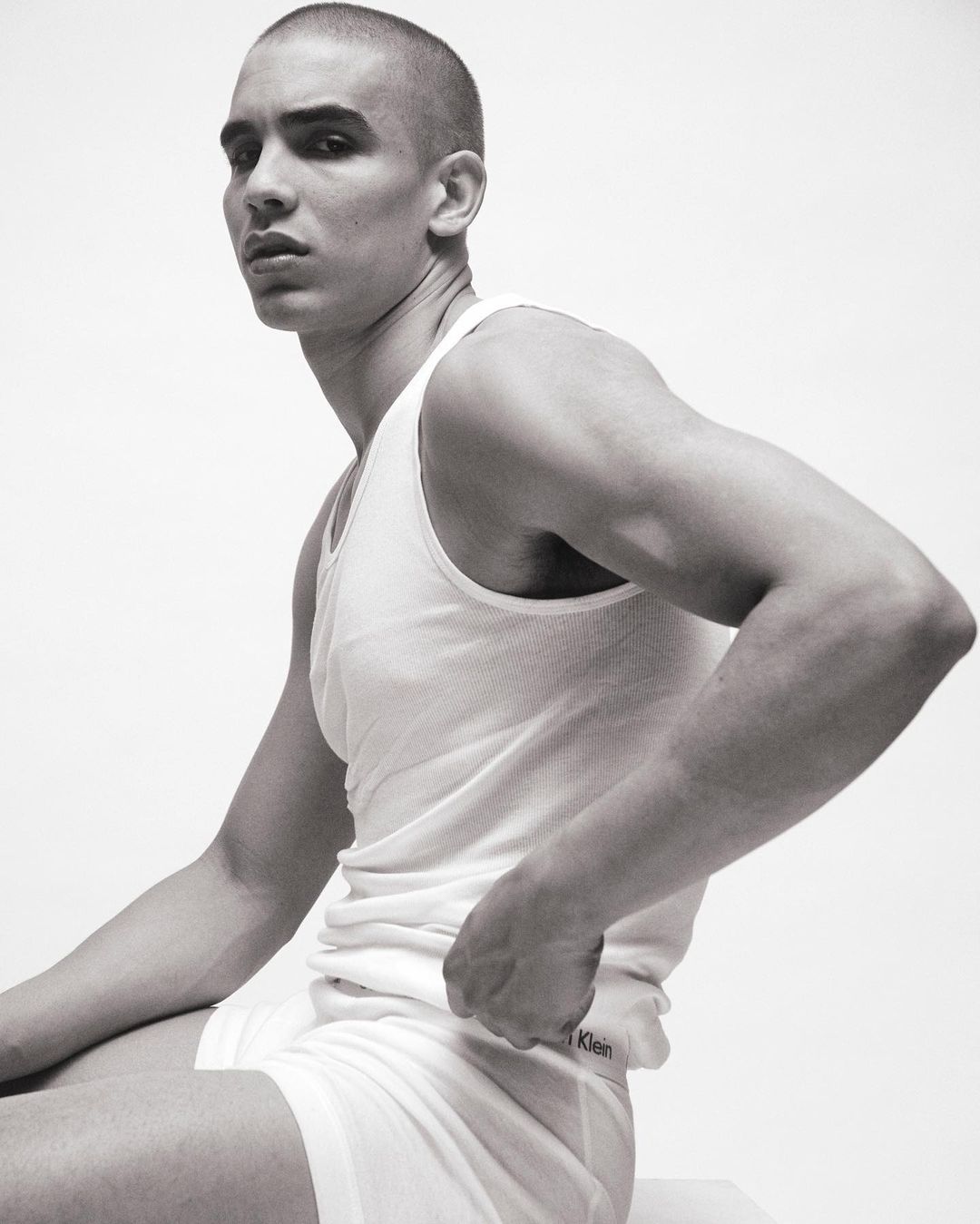 A man with a shaved head sits against a white background in underwear and a tank top, showcasing a minimalist fashion aesthetic.